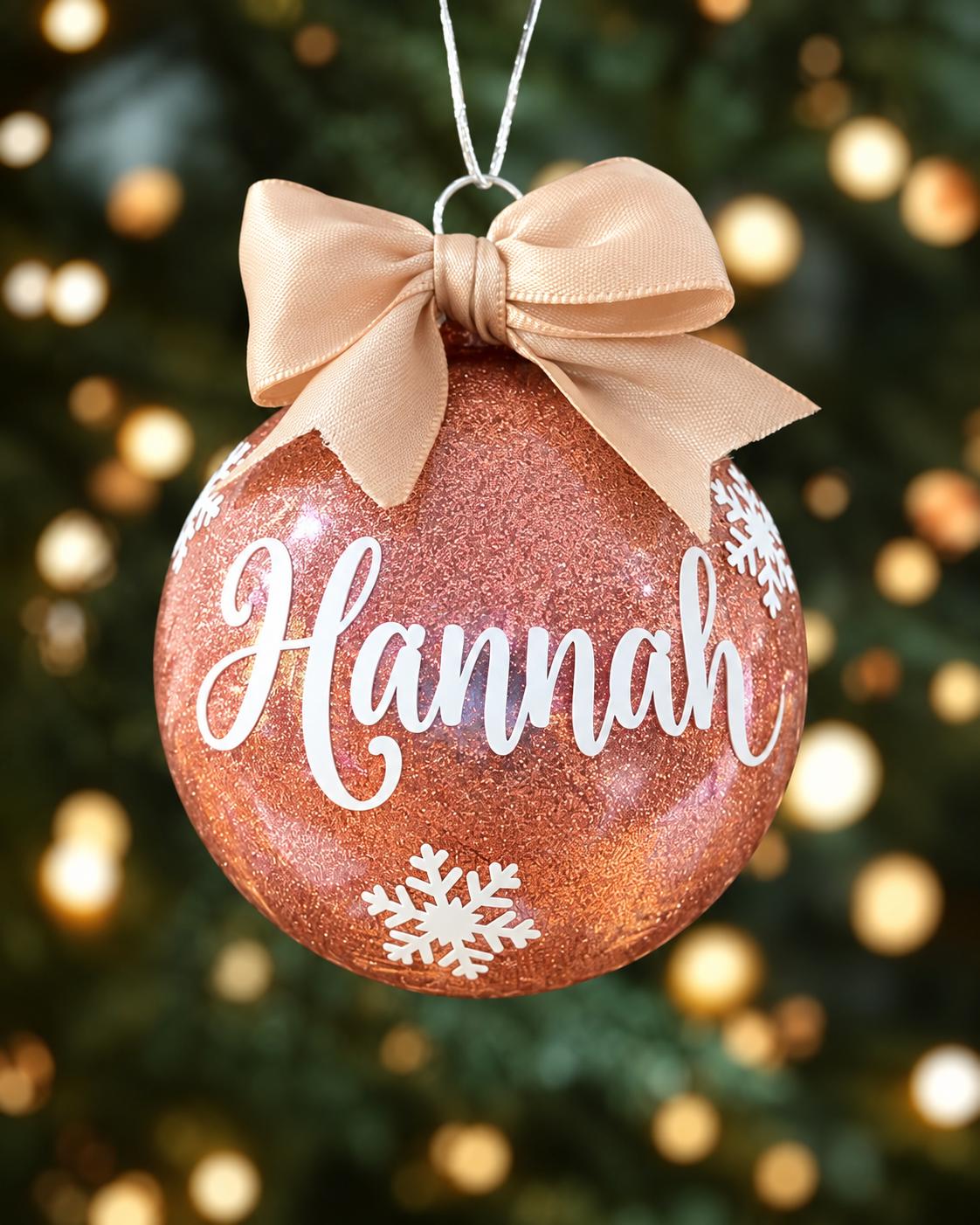 Rose gold glitter personalised name Christmas bauble reading Hannah with white snowflakes and a matching satin ribbon bow